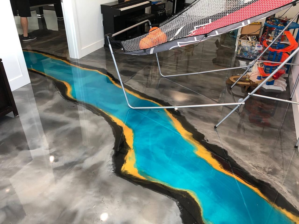A basement game room with a blue epoxy river flowing through it.  The blue epoxy river is flanked on both sides with yellow, black and swirled gray epoxy.