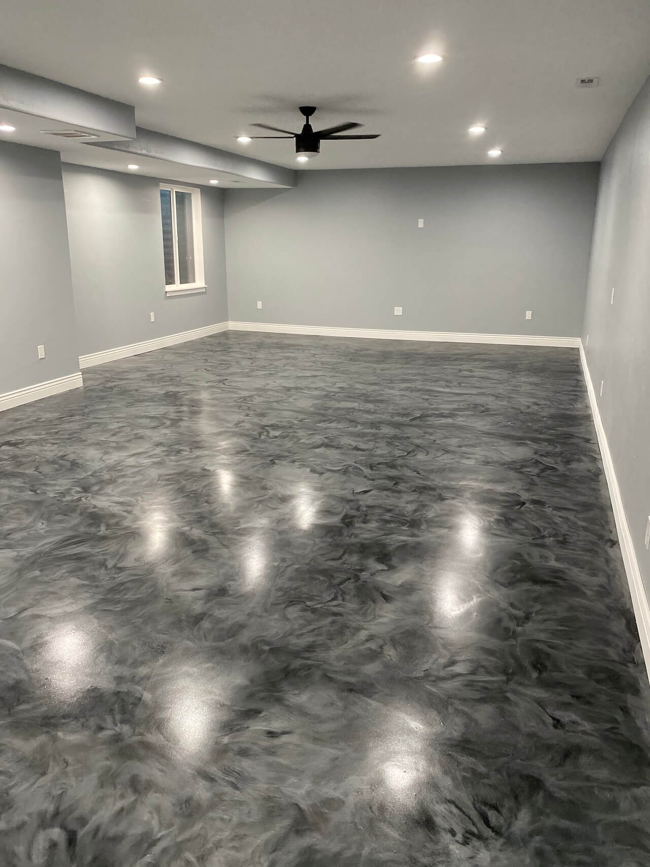 A basement that has been remodeled with Leggari epoxy floors in gray and black, new white trim, and freshly painted gray walls.