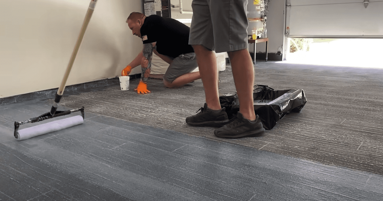 Applying the urethane gloss top coat to the hardwood overlay floor.  One person is applying it with a roller and another is applying it with a paint brush.