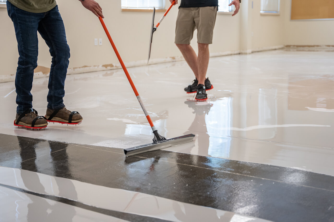 Two contractors wearing spike shoes using squeegees to smooth metallic epoxy out on a floor.