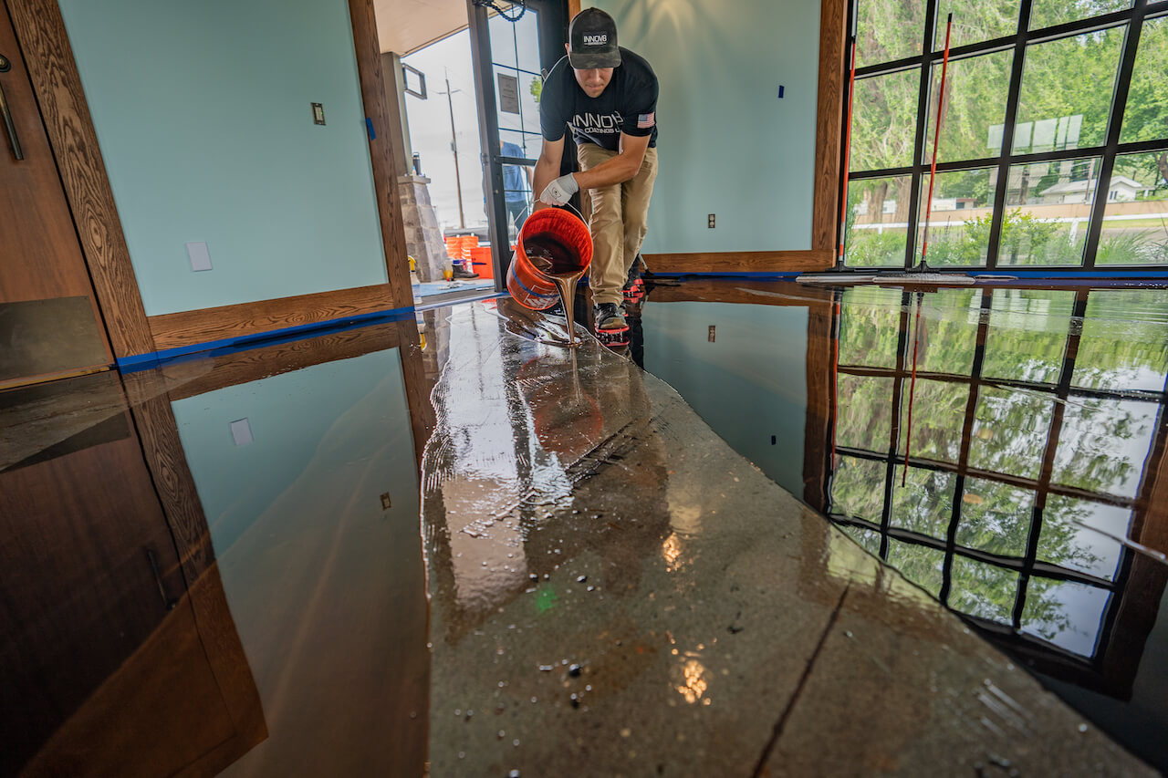 A contractor is applying Leggari metallic epoxy by pouring the mixed epoxy out of a bucket while wearing spike shoes.